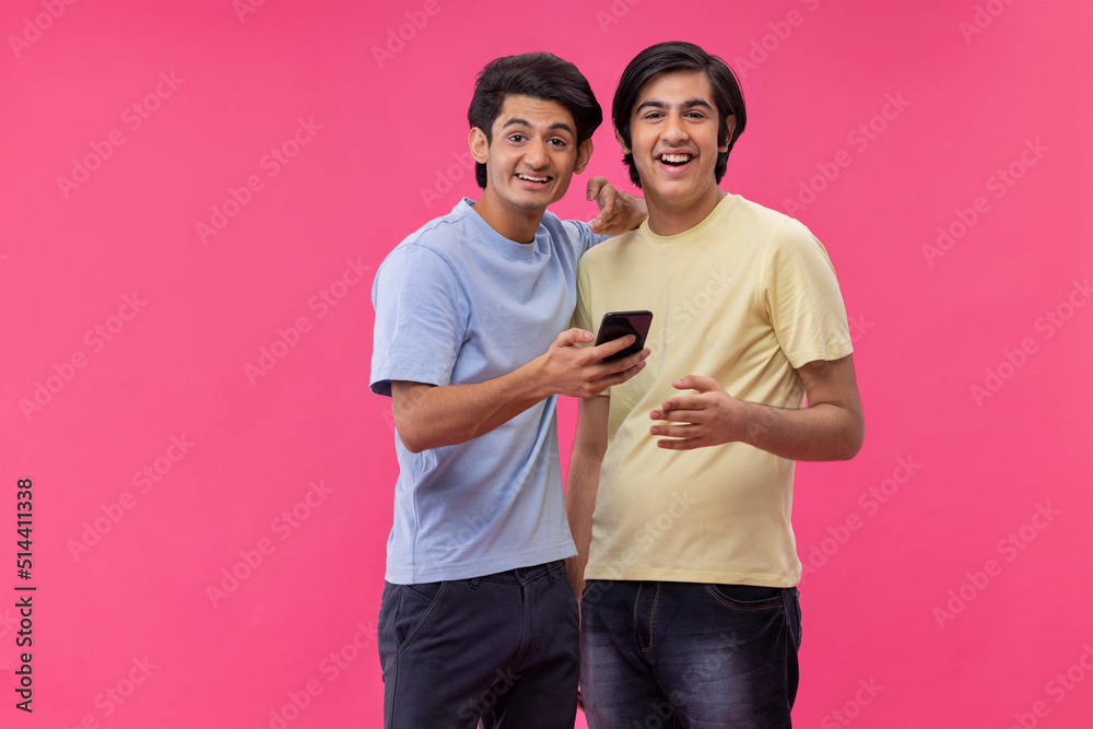 Two happy friends using smartphone while standing against pink background