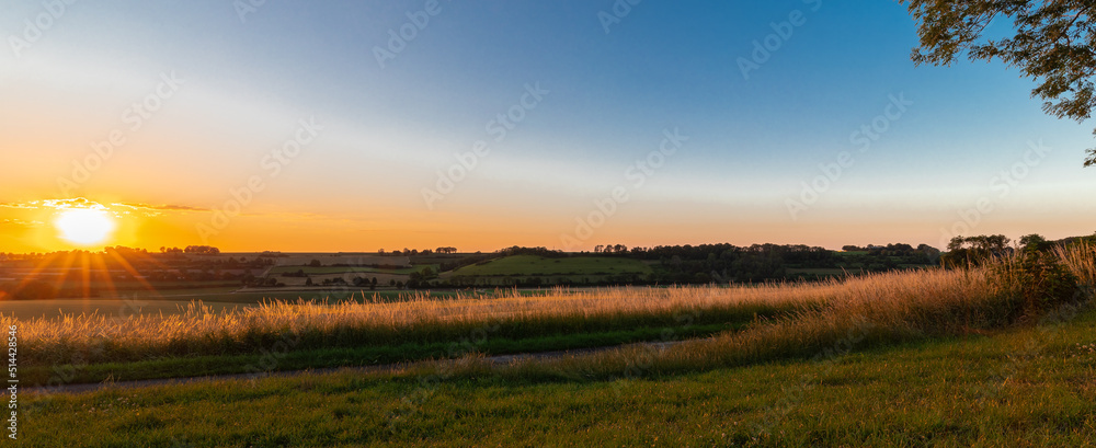 Fototapeta premium Sunset over the rolling hills in Elkenrade in the of south Limburg in the Netherlands with a spectacular view over the fields, full of wheat and some amazing beams from the sun during golden hour.