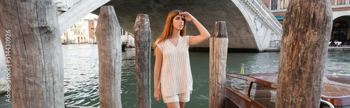 young woman in sleeveless jumper and shorts looking away near wooden pilings in Venice, banner.