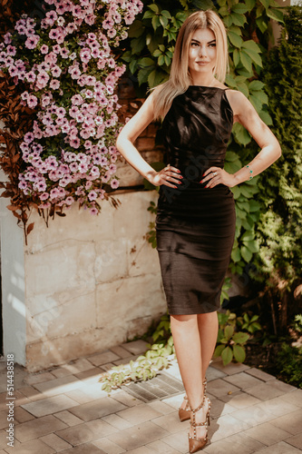 A girl in a dress with bare shoulders stands next to a bush with pink flowers. A girl walks through the streets of Spain.