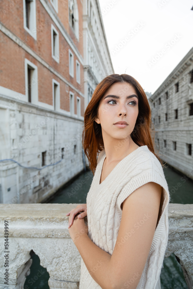 young woman in sleeveless jumper looking away near blurred venetian buildings.
