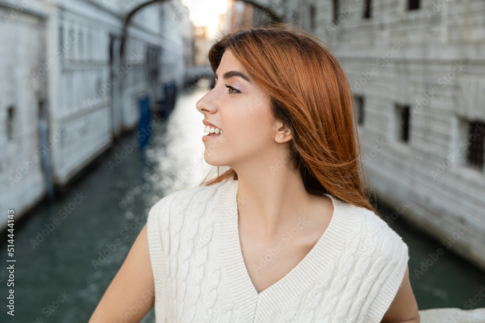 young and happy woman with red hair looking away on blurred background in Venice.