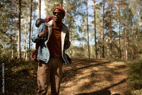 Curious african american tourist guy wander among trees with backpack in hat and sunglasses, happy man enjoying time alone in green forest. Escape from hectic life to wildlife. Search for adventures © Anatoliy Karlyuk