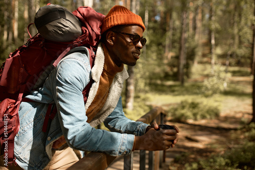 Happy african american guy in sunglasses and denim coat having rest leaning on wooden bridge in national park, admiring beauty of wild nature,with backpack on shoulders, holding thermos mug of tea