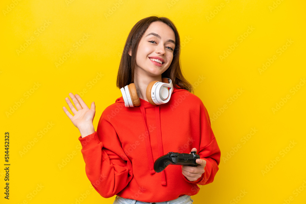 Young Ukrainian woman playing with a video game controller isolated on yellow background saluting with hand with happy expression