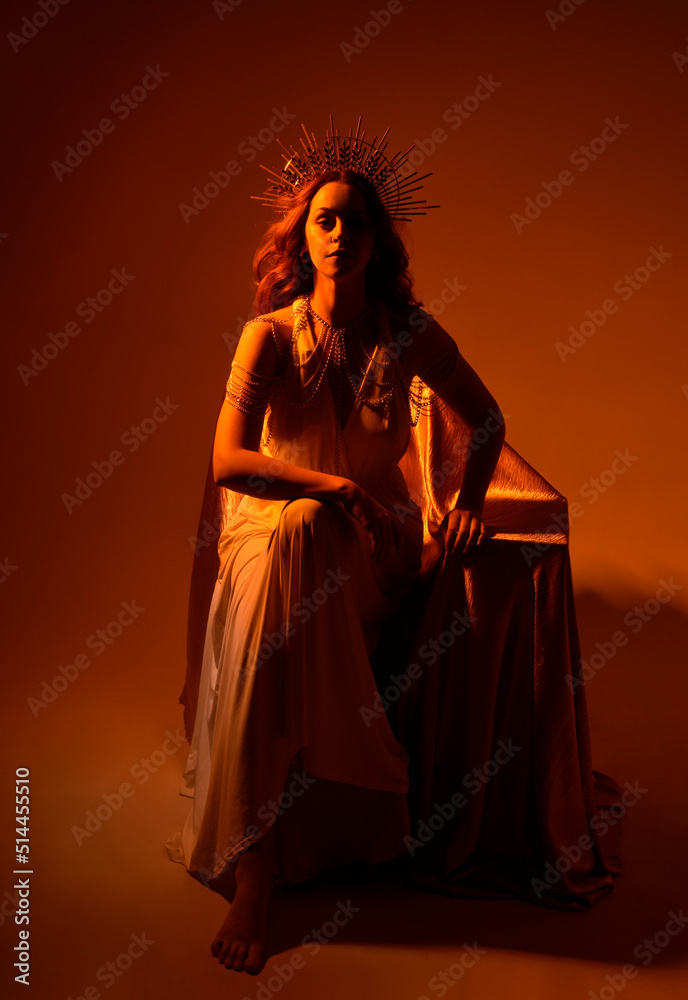 Full length portrait of beautiful red head woman wearing long flowing fantasy toga gown with golden halo crown jewellery,  sitting pose  a dark moody background with glowing or