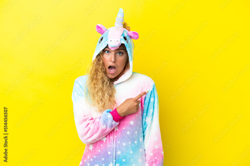 Girl with curly hair wearing a unicorn pajama isolated on yellow background surprised and pointing side Girl with curly hair wearing a unicorn pajama isolated on yellow background surprised and pointing side