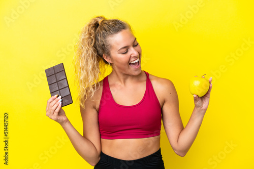 Girl with curly hair isolated on yellow background taking a chocolate tablet in one hand and an apple in the other