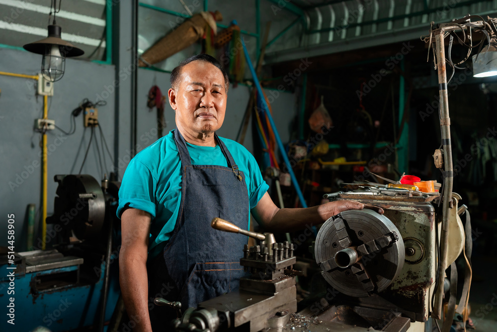 A senior Asian craftsman, standing closely and carefully controlling a lathe, inside a small factory of his family business..