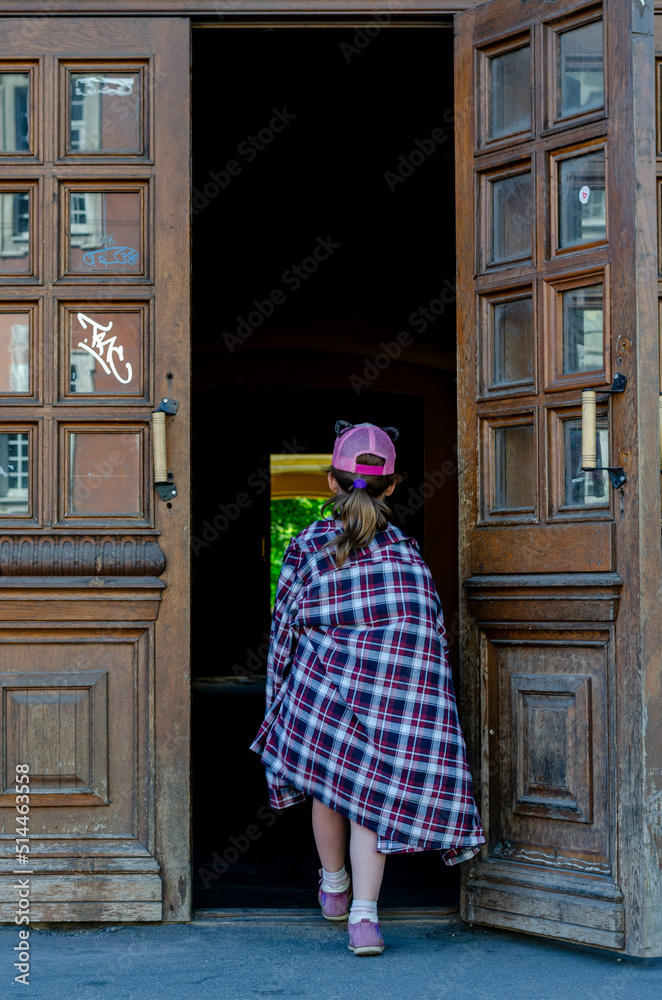 a girl in a pink cap and a large plaid cowboy shirt draped over her shoulders enters the doorway with her back to the camera through large beautiful doors with graffiti