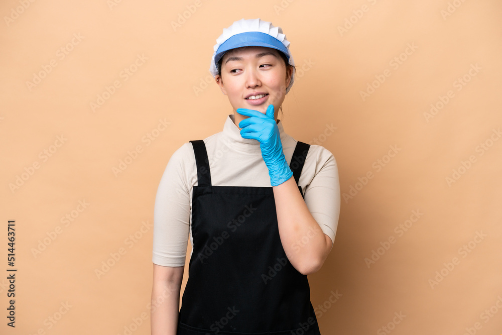 Young Chinese Fishmonger woman wearing an apron and holding a raw fish isolated on pink background looking to the side and smiling Young Chinese Fishmonger woman wearing an apron and holding a raw fish isolated on pink background looking to the side and smiling
