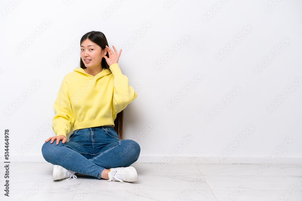 Young Chinese woman sitting on the floor isolated on white wall listening to something by putting hand on the ear Young Chinese woman sitting on the floor isolated on white wall listening to something by putting hand on the ear