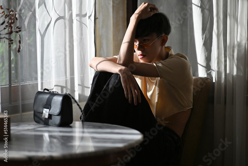 Portrait of lonely Asian gay teenager with crop top shirt and orange glasses sitting with knees up on chair, Lifestyle fashion LGBTQ concept. photo