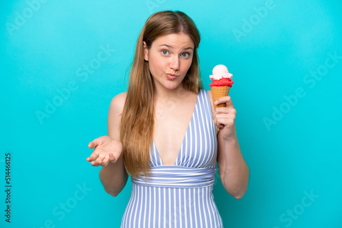 Young caucasian woman in swimsuit eating ice cream isolated on blue background making doubts gesture while lifting the shoulders