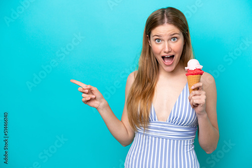 Young caucasian woman in swimsuit eating ice cream isolated on blue background surprised and pointing finger to the side
