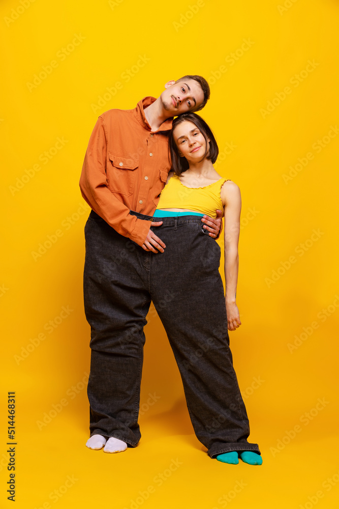 Obraz premium Creative colorful portrait of man and woman in bright clothes, standing together in one pants, hugging isolated over yellow background.