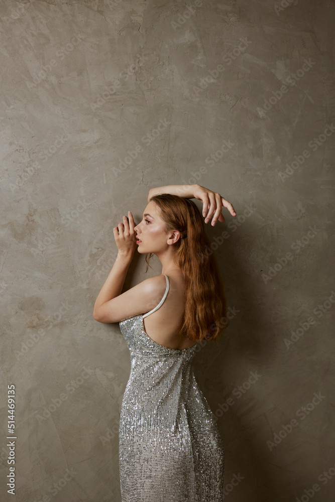 a gorgeous slender woman with long red hair poses standing in a shiny silver dress and straightening her hair with her hands a gorgeous slender woman with long red hair poses standing in a shiny silver dress and straightening her hair with her hands