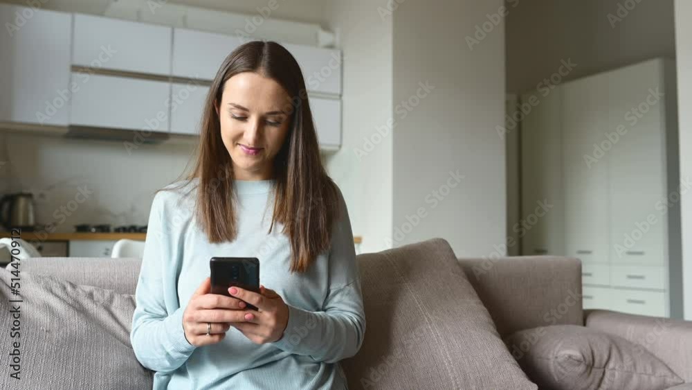 Serene young woman holds smartphone sitting on the couch at home, chatting online with friends, spend leisure time in social media, texting, scrolling news feed, shopping online Serene young woman holds smartphone sitting on the couch at home, chatting online with friends, spend leisure time in social media, texting, scrolling news feed, shopping online