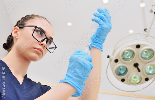 a pretty young woman in a surgical suit and glasses with a syringe in her hands against the background of a surgical lamp.