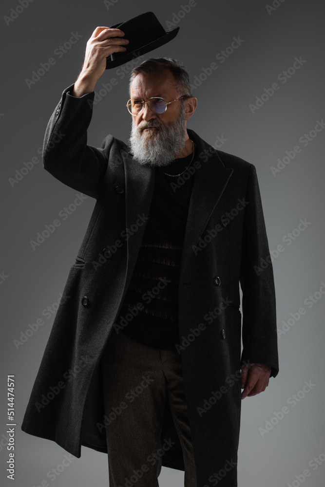 elegant senior man in stylish coat and eyeglasses wearing derby hat on grey. elegant senior man in stylish coat and eyeglasses wearing derby hat on grey.