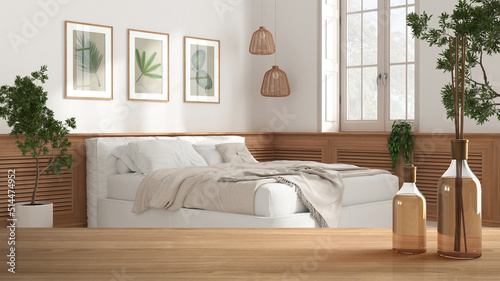 Wooden table top or shelf with aromatic sticks bottles over blurred bedroom with double soft bed, wooden wall panel, country provencal white architecture interior design