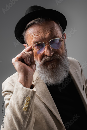 stylish senior man in beige jacket and derby hat adjusting eyeglasses on grey.