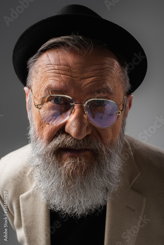 portrait of stylish senior man in eyeglasses and derby hat posing on grey.