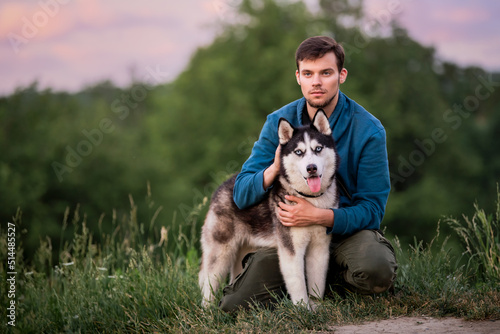 Portrait of a handsome young man and his pet Siberian Husky dog in nature in evening at sunset.