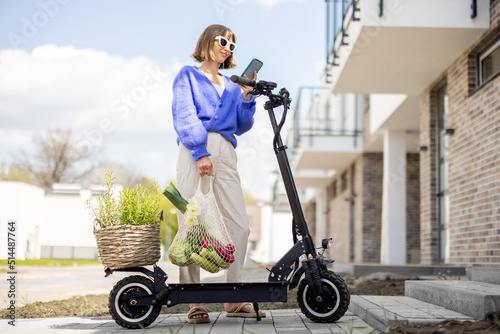 Happy stylish woman using phone while stannding with fresh vegetables and greens near electrical scooter at residential district. Concept of modern eco-friendly lifestyle and sustainability photo