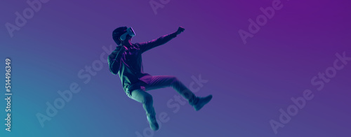 Young Asian man wearing VR headset playing video game and levitating in the air on futuristic purple cyberpunk neon light banner background. Metaverse technology concept.