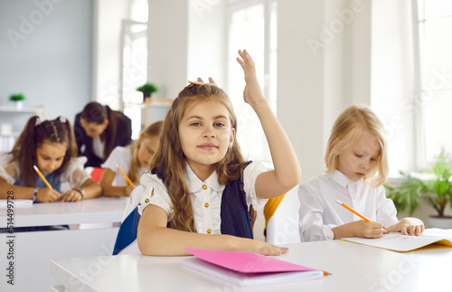 School student raising hand in class. Portrait of happy pretty student girl sitting at desk with notebook in classroom and raising up her hand. Back to school, elementary education, learning concept