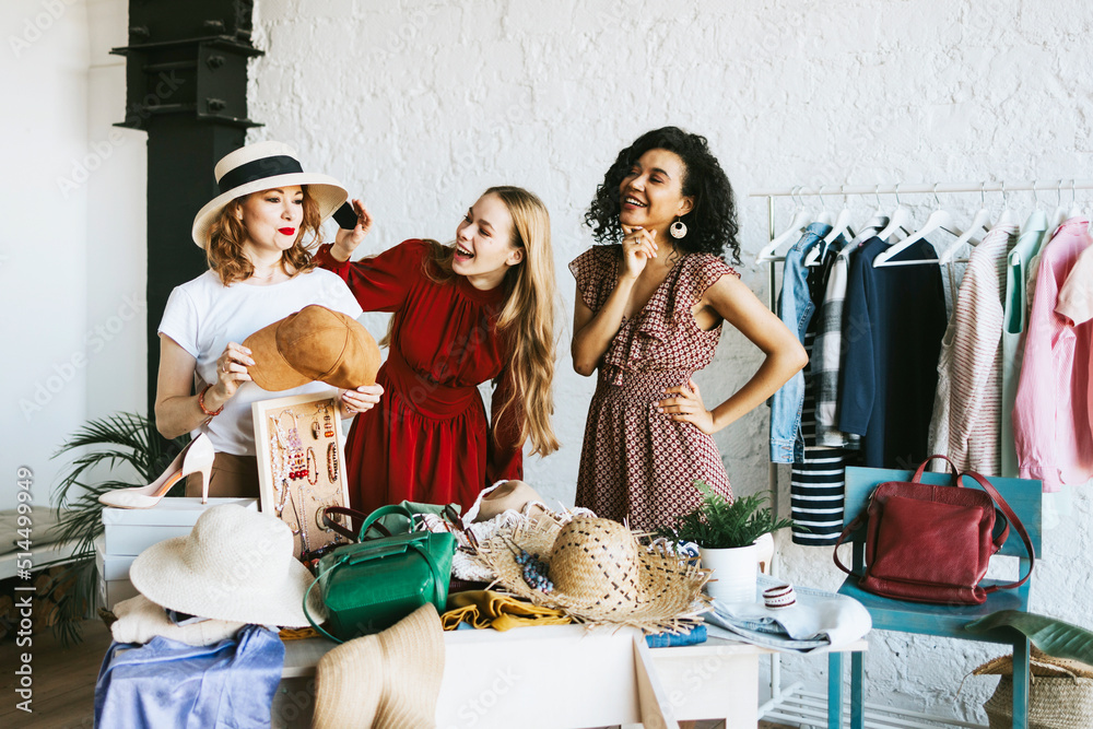 Obraz premium three young woman female caucasian and african students at swap party try on clothes, bags, shoes and accessories, change clothes with each other, second hand for things, zero waste life, eco-friendly