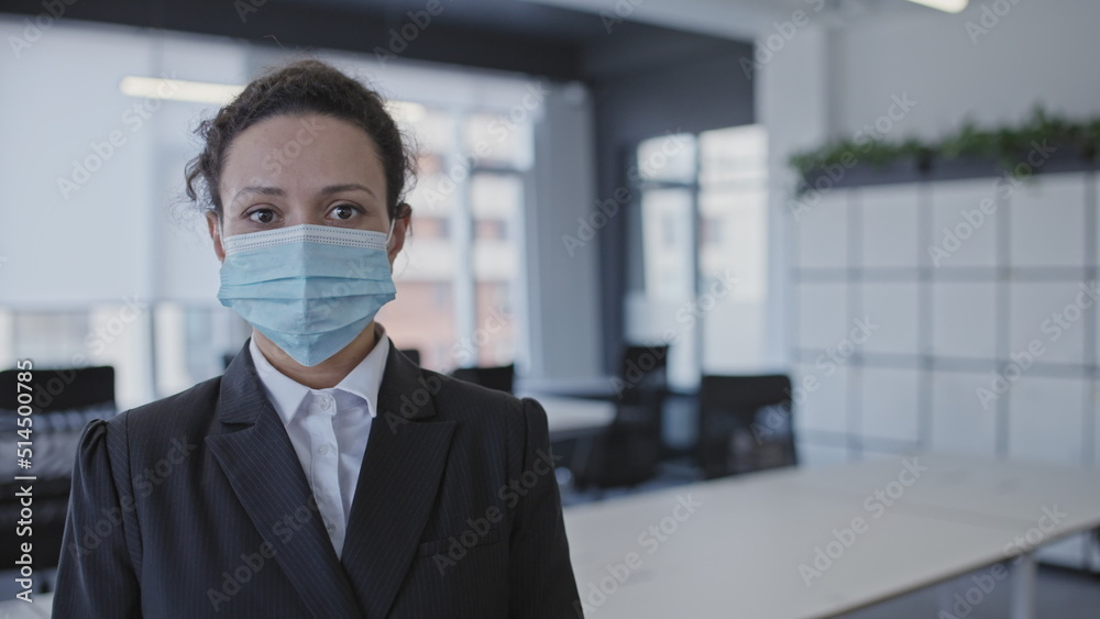 Businesswoman standing in empty office wearing protective face mask, coronavirus lockdown