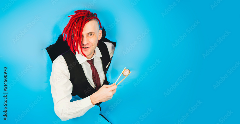Man with red dreadlocks holding in hand sticks chopsticks with sushi roll traditional japanese food looking at camera through hole in blue wall in studio Man with red dreadlocks holding in hand sticks chopsticks with sushi roll traditional japanese food looking at camera through hole in blue wall in studio