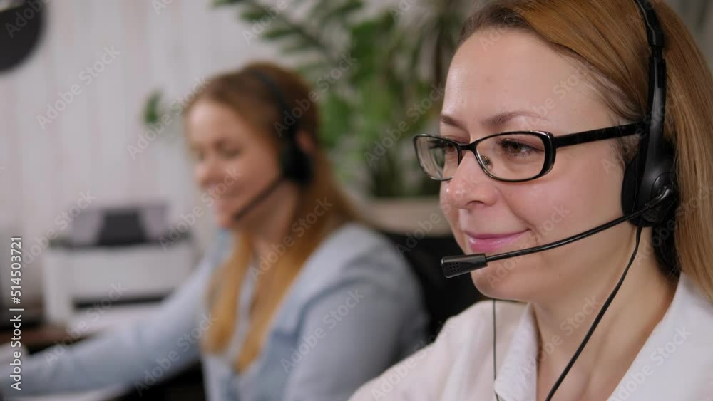 Close-up of a smiling woman in glasses with headphones working in a call center, she is talking to a client and looking at the computer screen. Close-up of a smiling woman in glasses with headphones working in a call center, she is talking to a client and looking at the computer screen.