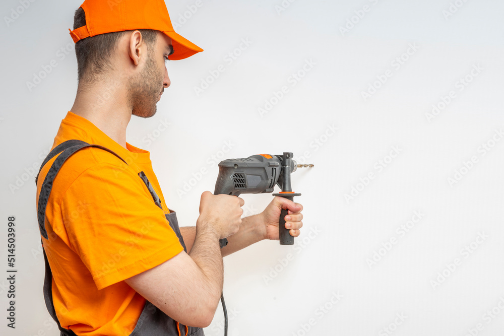 Young worker drilling with electric drill in his hands. Tool for boring holes. Craftsman repairman wearing orange uniform cap and t-shirt, overalls Young worker drilling with electric drill in his hands. Tool for boring holes. Craftsman repairman wearing orange uniform cap and t-shirt, overalls