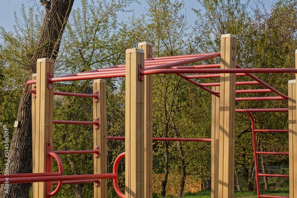 a sports ground with a red metal horizontal bar and an iron ladder on brown wooden poles on the street against the background of the sky and green vegetation a sports ground with a red metal horizontal bar and an iron ladder on brown wooden poles on the street against the background of the sky and green vegetation
