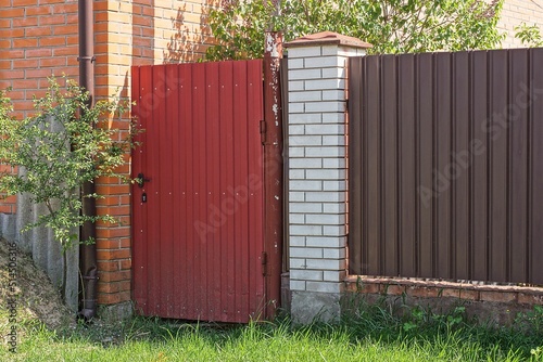 one red closed metal door on a fence wall made of white bricks and brown iron on a rural street in green grass