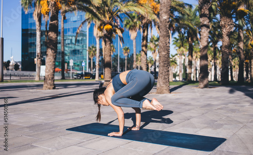 Strong female acrobat with muscular body doing frog handstand exercise during leisure at street with tropical trees, Asian woman dressed in active sportswear practicing balance asanas during warm up