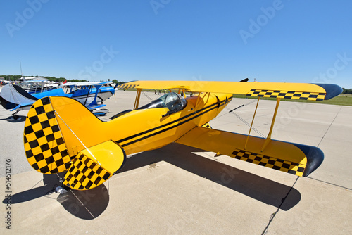 A small stunt biplane sits on the tarmac at an airshow under a brilliant sky.