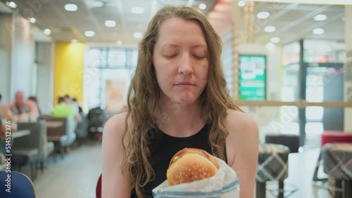 Woman with burger in a fast food restaurant. Woman opening wrapping paper, holding, biting the burger and savouring food. Fast snack on the go