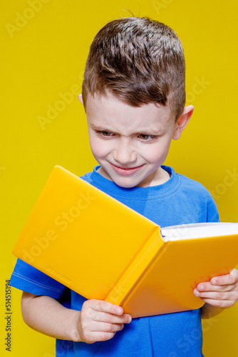 Little cheerful blond green-eyed boy 5-6 years old in a stylish blue T-shirt holding book and reading on yellow wall background, children's studio portrait