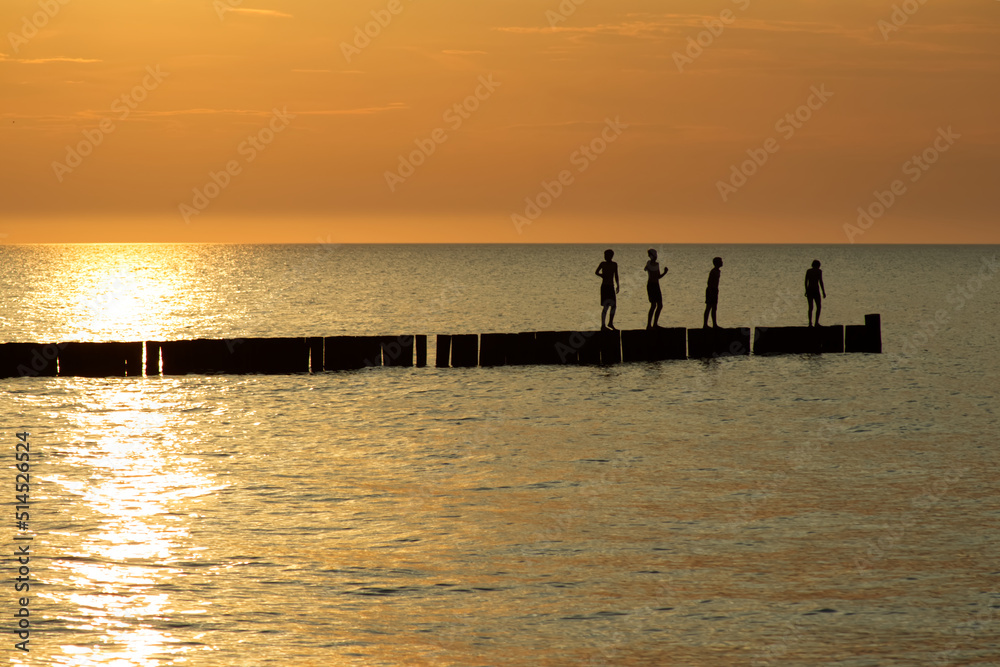 Four silhouettes of teenage children stand at breakwaters and at an orange sunset