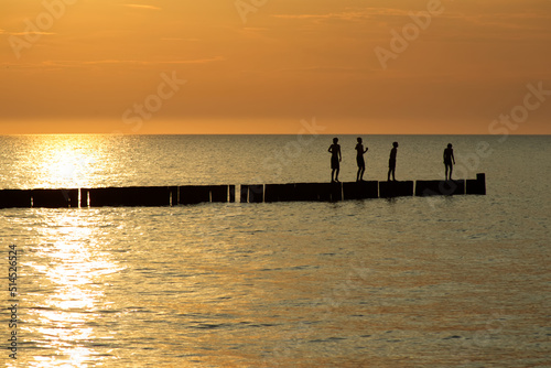 Four silhouettes of teenage children stand at breakwaters and at an orange sunset