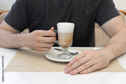 A young man in a cafe at the table holds a glass with latte coffee. A transparent cup of coffee on a saucer with a spoon in beautiful hands.