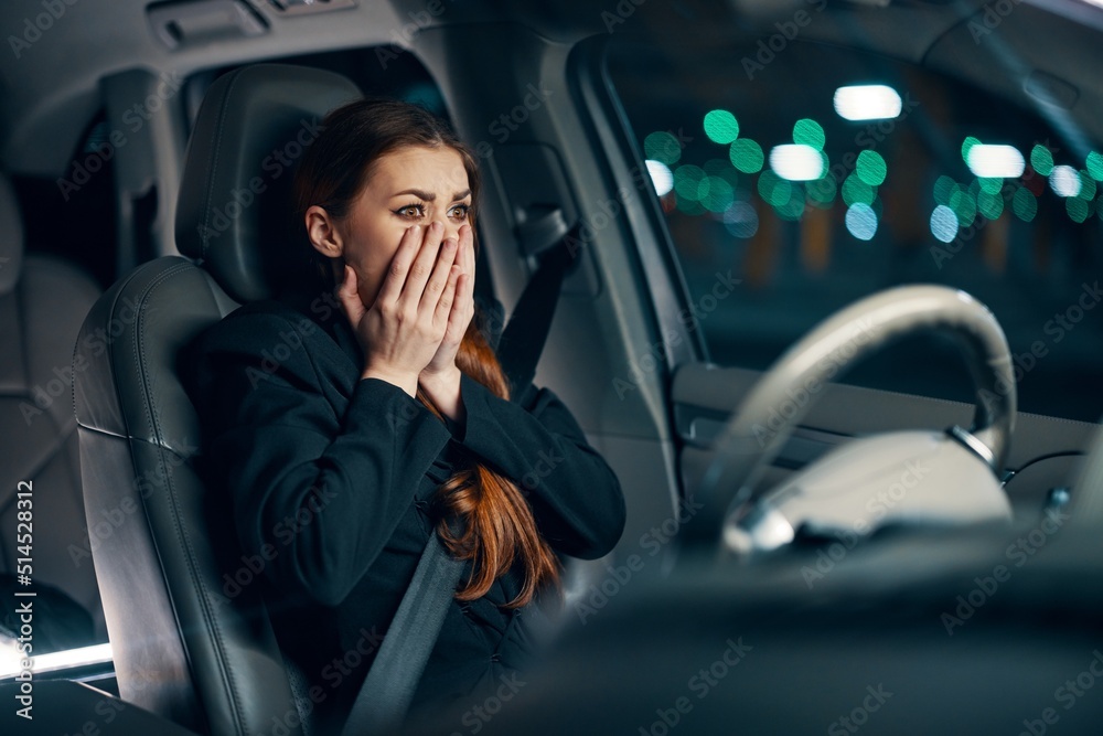 an emotional, frightened woman is sitting behind the wheel of a car in a black shirt, wearing a seat belt, expressing her emotions, covering her face with her hands. Photography at night 