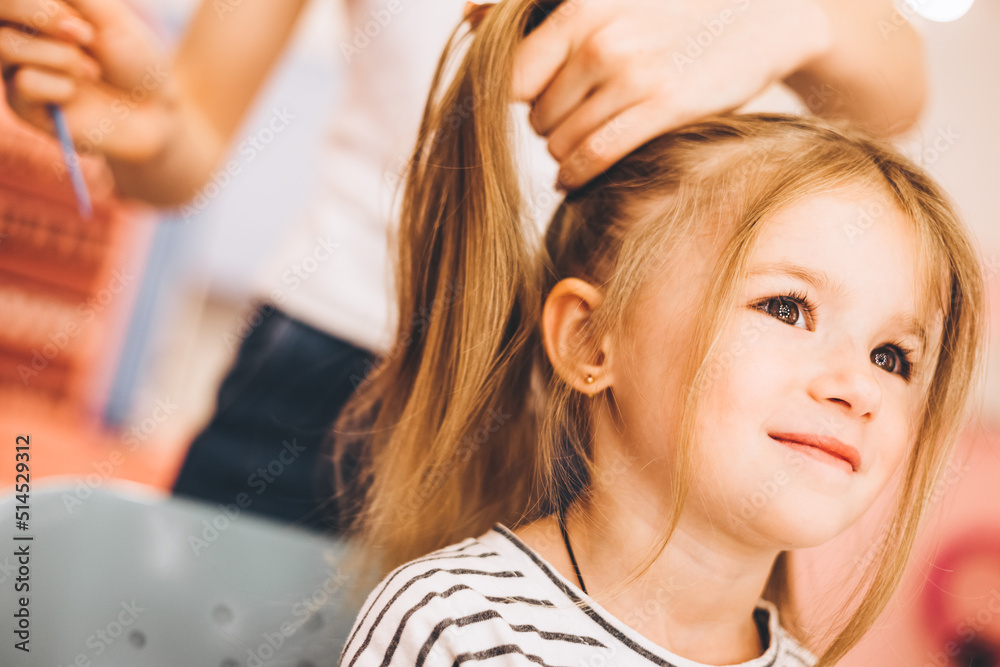 Naklejka premium Stylish little girl in a beauty salon for children where she made a beautiful hairstyle. Female stylist makes stylish hairstyle to little girl.
