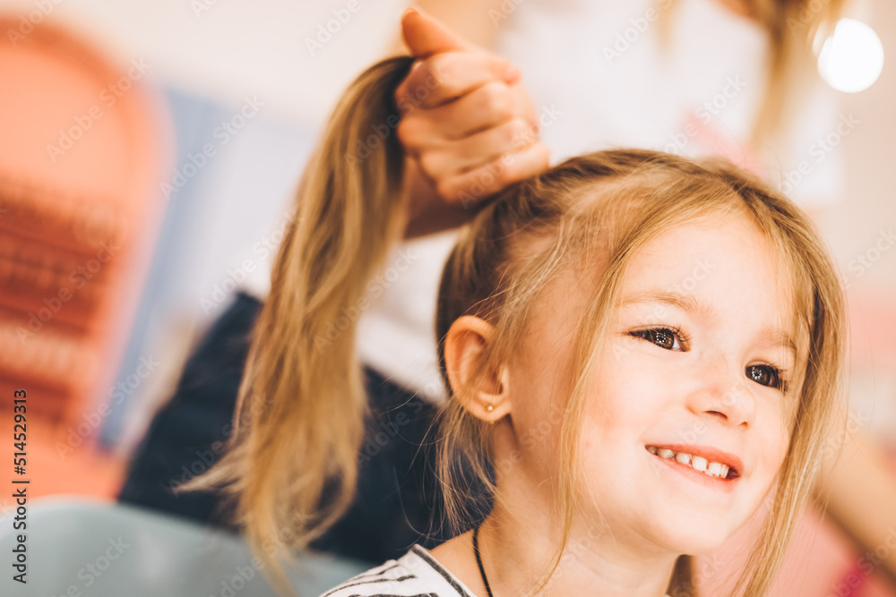 Fototapeta premium Stylish little girl in a beauty salon for children where she made a beautiful hairstyle. Female stylist makes stylish hairstyle to little girl.