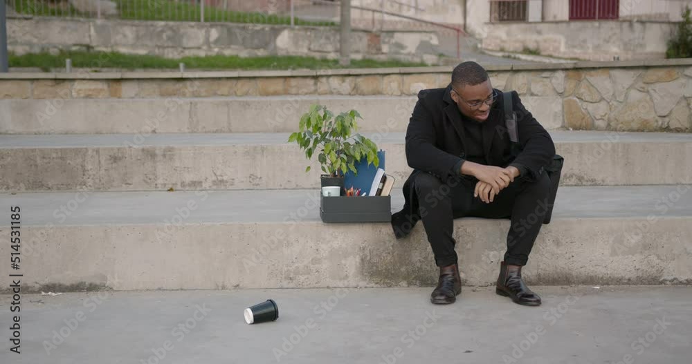 Disappointed black employee with box of office stuff sitting on stairs Disappointed black employee with box of office stuff sitting on stairs
