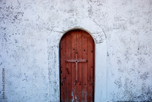 Wooden door of a hermitage in the town "La Peral" in Somiedo, Asturias, Spain. The doorway has a nailed wooden cross and arched top and is surrounded by an old cracked wall painted white © AntonioLopez